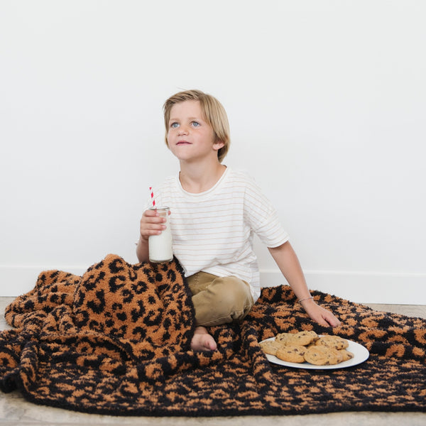 A little boy has cookies and milk and is with a luxurious double-layer Bamboni® blanket by Saranoni, featuring classic leopard print of orange and black. This ultra-soft blanket is crafted from bamboo rayon and poly microfiber, making it lightweight, breathable, and fluffy. Ideal as a cozy blanket, extra large blanket, luxury blanket, soft blanket, or stylish home accent, this Saranoni blanket brings warmth, comfort, and timeless elegance to any living room or bedroom.