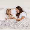 A mother and daughter sit with a luxurious double-layer Bamboni® blanket by Saranoni, featuring gray leopard print that is white and gray. This ultra-soft blanket is crafted from bamboo rayon and poly microfiber, making it lightweight, breathable, and fluffy. Ideal as a cozy blanket, extra large blanket, luxury blanket, soft blanket, or stylish home accent, this Saranoni blanket brings warmth, comfort, and timeless elegance to any living room or bedroom.