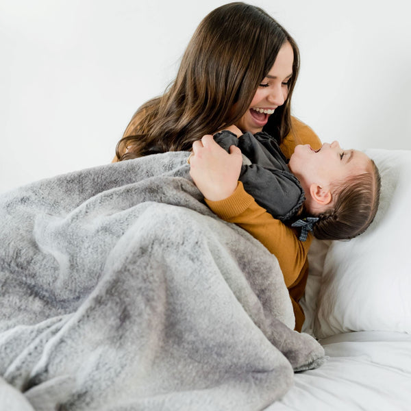 A mother and her little girl with a Saranoni Grand Faux Fur blanket, featuring an elegant white and gray and brown tone and luxuriously soft texture. This cozy, throw blanket and faux fur blanket offers soothing weight, premium softness, timeless comfort, and is a soft blanket. Perfect as a cozy blanket and luxury blanket for snuggling or as a luxury bedroom accent, this Saranoni blanket elevates every cozy moment and is a cozy blanket.