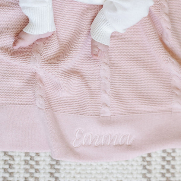 A close-up of a baby resting on a soft, pink Saranoni Heirloom Cotton Knit Receiving Blanket. The luxury 100% cotton blanket features classic cable knit detailing, a wide border, and the name "Emma" embroidered on it, making it a personalized and timeless keepsake. The baby’s hands rest on the cozy, elevated blanket, perfect for special occasions like baby showers or newborn photoshoots. Ideal for searches related to personalized baby blankets, soft cotton receiving blankets, or heirloom-quality baby gifts.