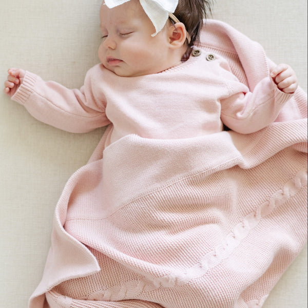 A close-up of a baby resting on a soft, pink Saranoni Heirloom Cotton Knit Receiving Blanket. The luxury 100% cotton blanket features classic cable knit detailing, a wide border, making it a timeless keepsake. The cozy, elevated blanket, is perfect for special occasions like baby showers or newborn photoshoots. Ideal for searches related to personalized baby blankets, soft cotton receiving blankets, or heirloom-quality baby gifts.