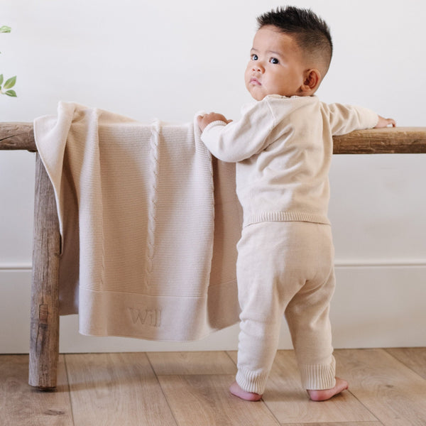 A baby boy wearing a tan Saranoni Cotton Set stands by a soft, tan Saranoni Heirloom Cotton Knit Receiving Blanket. The luxury 100% cotton blanket features classic cable knit detailing, a wide border, making it a timeless keepsake. The cozy, elevated blanket, is perfect for special occasions like baby showers or newborn photoshoots. Ideal for searches related to personalized baby blankets, soft cotton receiving blankets, or heirloom-quality baby gifts.