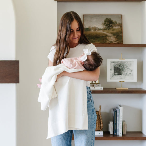 A mother holds her baby girl in a soft, white Saranoni Heirloom Cotton Knit Receiving Blanket. The luxury 100% cotton blanket features classic cable knit detailing, a wide border, making it a timeless keepsake. The cozy, elevated blanket, is perfect for special occasions like baby showers or newborn photoshoots. Ideal for searches related to personalized baby blankets, soft cotton receiving blankets, or heirloom-quality baby gifts.