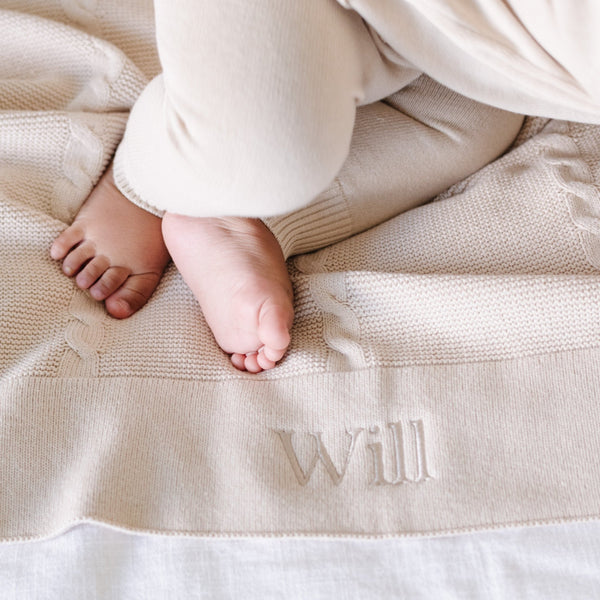 A baby boy lays on a soft, tan Saranoni Heirloom Cotton Knit Receiving Blanket with the name "Will" embroidered on it. The luxury 100% cotton blanket features classic cable knit detailing, a wide border, making it a timeless keepsake. The cozy, elevated blanket, is perfect for special occasions like baby showers or newborn photoshoots. Ideal for searches related to personalized baby blankets, soft cotton receiving blankets, or heirloom-quality baby gifts.