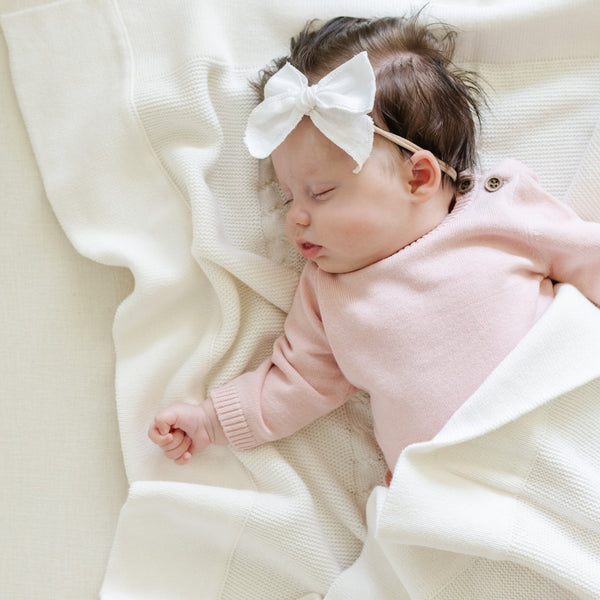 A baby girl in a Saranoni Heirloom Cotton Knit Set lays on a soft, white Saranoni Heirloom Cotton Knit Receiving Blanket. The luxury 100% cotton blanket features classic cable knit detailing, a wide border, making it a timeless keepsake. The cozy, elevated blanket, is perfect for special occasions like baby showers or newborn photoshoots. Ideal for searches related to personalized baby blankets, soft cotton receiving blankets, or heirloom-quality baby gifts.