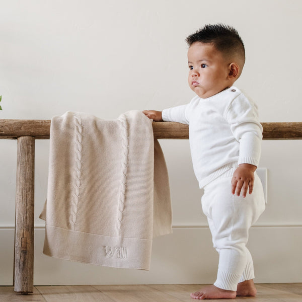 A baby boy wearing a white Saranoni Cotton Set stands by a soft, tan Saranoni Heirloom Cotton Knit Receiving Blanket. The luxury 100% cotton blanket features classic cable knit detailing, a wide border, making it a timeless keepsake. The cozy, elevated blanket, is perfect for special occasions like baby showers or newborn photoshoots. Ideal for searches related to personalized baby blankets, soft cotton receiving blankets, or heirloom-quality baby gifts.