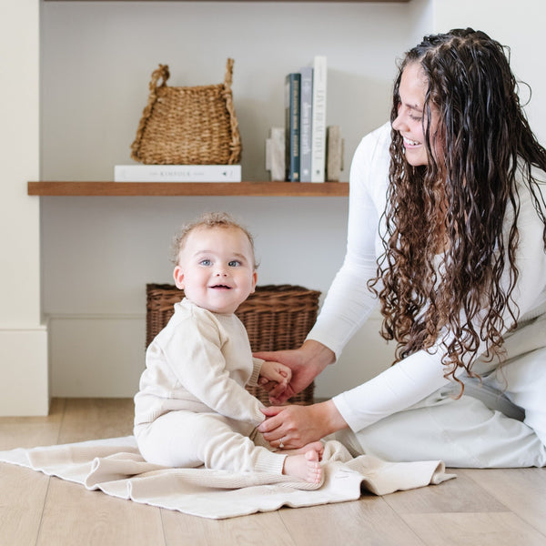 A mother plays with her baby on a soft, tan Saranoni Heirloom Cotton Knit Receiving Blanket. The luxury 100% cotton blanket features classic cable knit detailing, a wide border, making it a timeless keepsake. The cozy, elevated blanket, is perfect for special occasions like baby showers or newborn photoshoots. Ideal for searches related to personalized baby blankets, soft cotton receiving blankets, or heirloom-quality baby gifts.