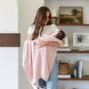 A mother holds her baby in a soft, pink Saranoni Heirloom Cotton Knit Receiving Blanket. The luxury 100% cotton blanket features classic cable knit detailing, a wide border, making it a timeless keepsake. The cozy, elevated blanket, is perfect for special occasions like baby showers or newborn photoshoots. Ideal for searches related to personalized baby blankets, soft cotton receiving blankets, or heirloom-quality baby gifts.