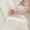 A baby girl lays on a soft, white Saranoni Heirloom Cotton Knit Receiving Blanket. The luxury 100% cotton blanket features classic cable knit detailing, a wide border, making it a timeless keepsake. The cozy, elevated blanket, is perfect for special occasions like baby showers or newborn photoshoots. Ideal for searches related to personalized baby blankets, soft cotton receiving blankets, or heirloom-quality baby gifts.