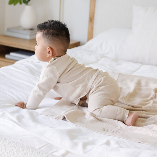 A baby boy crawls on a Saranoni Heirloom Knit Blanket. He is wearing a light tan, 100% cotton baby outfit that combines style and comfort. The Saranoni cloud-soft cotton set features a breathable waistband, loosely fitted sleeves, and elevated button details, making it perfect for everyday wear or special occasions. This heirloom-quality baby clothing offers timeless style and ultimate comfort. Ideal for parents searching for luxury baby clothes, soft cotton baby sets, or high-quality infant outfit.