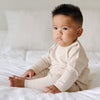 A baby boy sits on a bed. He is wearing a light tan, 100% cotton baby outfit that combines style and comfort. The Saranoni cloud-soft cotton set features a breathable waistband, loosely fitted sleeves, and elevated button details, making it perfect for everyday wear or special occasions. This heirloom-quality baby clothing offers timeless style and ultimate comfort. Ideal for parents searching for luxury baby clothes, soft cotton baby sets, or high-quality infant outfit.