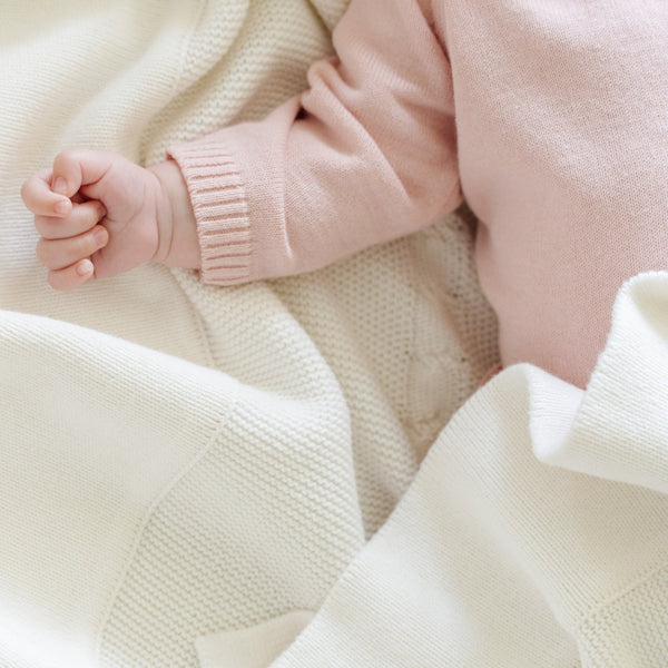 A baby girl lays on a Saranoni Heirloom Knit Blanket. She is wearing a light pink, 100% cotton baby outfit that combines style and comfort. The Saranoni cloud-soft cotton set features a breathable waistband, loosely fitted sleeves, and elevated button details, making it perfect for everyday wear or special occasions. This heirloom-quality baby clothing offers timeless style and ultimate comfort. Ideal for parents searching for luxury baby clothes, soft cotton baby sets, or high-quality infant outfit.