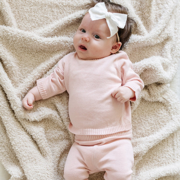 A baby girl lays on a Saranoni Blanket. She is wearing a light pink, 100% cotton Saranoni baby outfit that combines style and comfort. The Saranoni cloud-soft cotton set features a breathable waistband, loosely fitted sleeves, and elevated button details, making it perfect for everyday wear or special occasions. This heirloom-quality baby clothing offers timeless style and ultimate comfort. Ideal for parents searching for luxury baby clothes, soft cotton baby sets, or high-quality infant outfit.