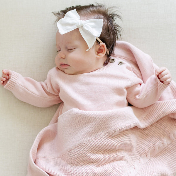 A baby girl lays on a Saranoni Heirloom Knit Blanket. She is wearing a light pink, 100% cotton baby outfit that combines style and comfort. The Saranoni cloud-soft cotton set features a breathable waistband, loosely fitted sleeves, and elevated button details, making it perfect for everyday wear or special occasions. This heirloom-quality baby clothing offers timeless style and ultimate comfort. Ideal for parents searching for luxury baby clothes, soft cotton baby sets, or high-quality infant outfit.