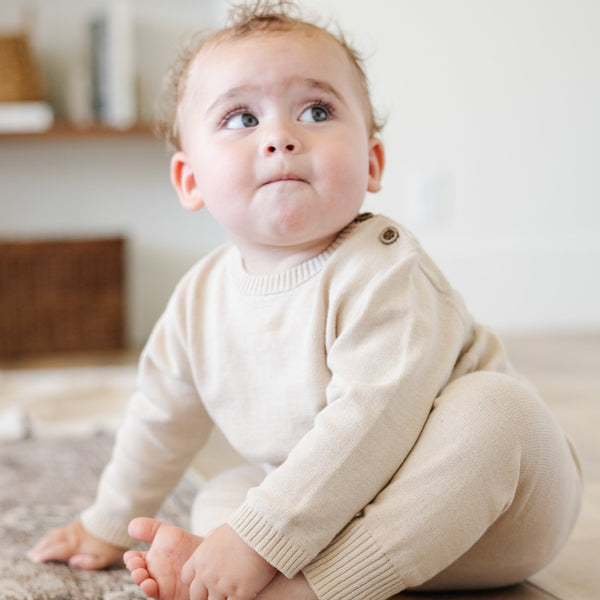 A baby boy sits on a wooden floor. He is wearing a light tan, 100% cotton baby outfit that combines style and comfort. The Saranoni cloud-soft cotton set features a breathable waistband, loosely fitted sleeves, and elevated button details, making it perfect for everyday wear or special occasions. This heirloom-quality baby clothing offers timeless style and ultimate comfort. Ideal for parents searching for luxury baby clothes, soft cotton baby sets, or high-quality infant outfit.