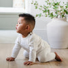 A baby boy is crawling on a wooden floor. He is wearing an all-white, 100% cotton baby outfit that combines style and comfort. The Saranoni cloud-soft cotton set features a breathable waistband, loosely fitted sleeves, and elevated button details, making it perfect for everyday wear or special occasions. This heirloom-quality baby clothing offers timeless style and ultimate comfort. Ideal for parents searching for luxury baby clothes, soft cotton baby sets, or high-quality infant outfit.