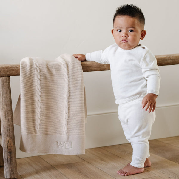 A baby boy stands stands by a Saranoni Heirloom Knit Blanket. He is wearing an all-white, 100% cotton baby outfit that combines style and comfort. The Saranoni cloud-soft cotton set features a breathable waistband, loosely fitted sleeves, and elevated button details, making it perfect for everyday wear or special occasions. This heirloom-quality baby clothing offers timeless style and ultimate comfort. Ideal for parents searching for luxury baby clothes, soft cotton baby sets, or high-quality infant outfit.