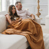 A husband and wife sit holding a Luxury Throw Camel (light yellowish brown) Colored Lush Saranoni Blanket. The soft blanket is a throw blanket and has a soft faux fur feeling.