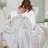 A mother and son sit holding a Luxury Throw Feather (light gray) Colored Lush Saranoni Blanket. The soft blanket is a throw blanket and has a soft faux fur feeling.