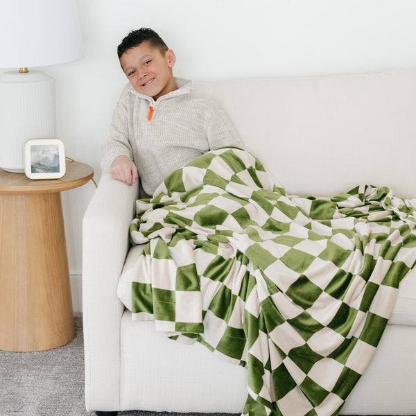 Boy sitting on couch wrapped in Olive green checkered MINKY STRETCH XL THROW BLANKETS - Saranoni