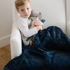 A little boy sits under a Luxury Extra Large Oversized Throw Navy Colored Lush Saranoni Blanket. The soft blanket is an extra large throw blanket and has a soft faux fur feeling.