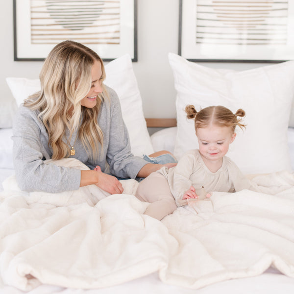 A mother and daughter sit with a Luxury Extra Large Oversized Throw Natural (off-white) Colored Lush Saranoni Blanket. The soft blanket is an extra large throw blanket and has a soft faux fur feeling.