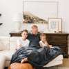 A father sits with his two sons under a Luxury Extra Large Oversized Throw Charcoal Colored Lush Saranoni Blanket. The soft blanket is an extra large throw blanket and has a soft faux fur feeling.