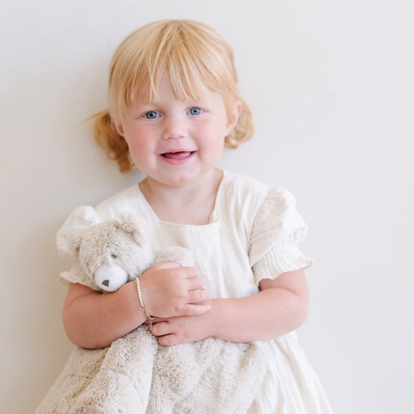 Adorable toddler girl in a white dress cuddling a Saranoni bear stuffed animal lovey. This ultra-soft plush lovey blanket features a cozy bear head and luxuriously soft fabric, designed to provide comfort, security, and soothing snuggles for babies and toddlers. Made with high-quality materials, the Saranoni lovey is perfect for newborns, infants, and young children as a bedtime companion or travel comfort item. Ideal baby shower gift or nursery essential, offering warmth and softness in a neutral design. 