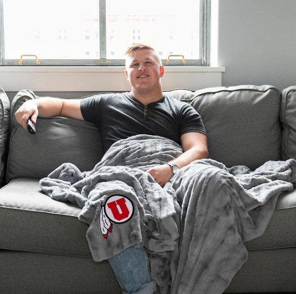 A man sits holding a Luxury Throw Gray Colored Patterned Faux Fur Saranoni Blanket. The soft blanket is a throw blanket and has a soft faux fur feeling. The blanket has a University of Utah Logo.