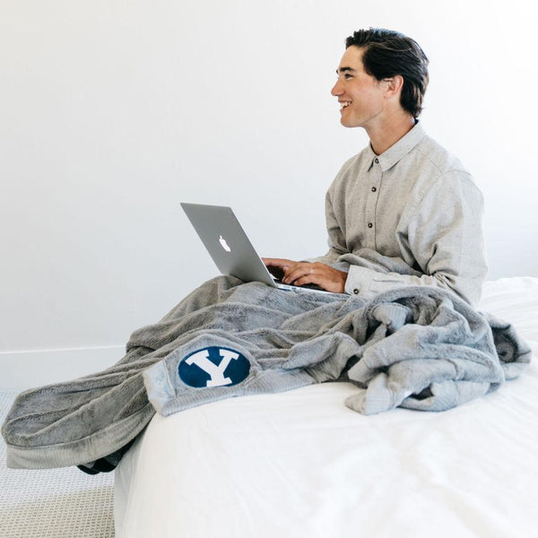 A boy sits with a Luxury Extra Large Oversized Throw Gray Colored Lush Saranoni Blanket. The soft blanket has a soft faux fur feeling. The extra large blanket has the Brigham Young University Logo on it.