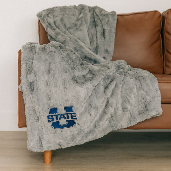 A Luxury Throw Gray Colored Patterned Faux Fur Saranoni Blanket. The soft blanket is a throw blanket and has a soft faux fur feeling. The blanket has a Utah State University Logo.