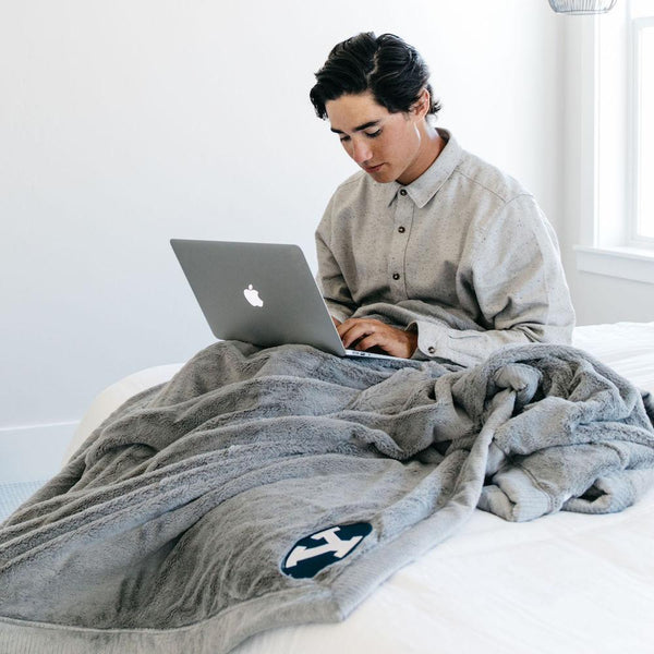 A boy sits with a Luxury Extra Large Oversized Throw Gray Colored Lush Saranoni Blanket. The soft blanket has a soft faux fur feeling. The extra large blanket has the Brigham Young University Logo on it.