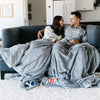 A husband and wife sit with two Luxury Extra Large Oversized Throw Gray Colored Lush Saranoni Blanket. The soft blanket has a soft faux fur feeling. One extra large blanket has the Brigham Young University Logo on it and the other has a University of Utah Logo.