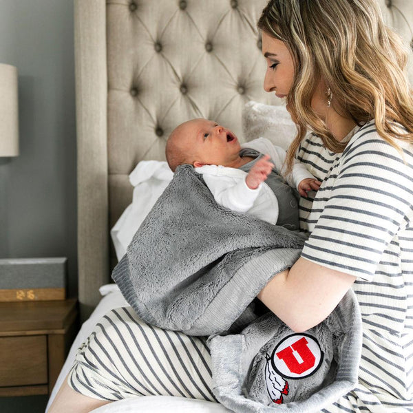 A mother holds her baby while holding a Luxury Receiving Gray Colored Lush Saranoni Blanket. The soft blanket is a toddler blanket or baby blanket and has a soft faux fur feeling. The blanket has the University of Utah Logo on it.