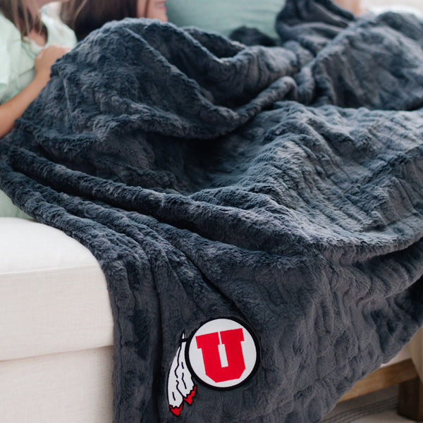 A Luxury Extra Large Throw Charcoal Colored Patterned Faux Fur Saranoni Blanket. The soft blanket is an oversized throw blanket and has a soft faux fur feeling. The blanket has a University of Utah Logo.