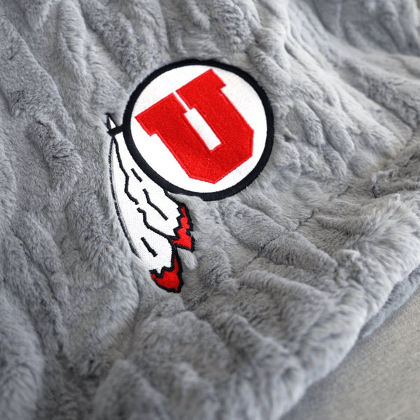 A Luxury Throw Gray Colored Patterned Faux Fur Saranoni Blanket. The soft blanket is a throw blanket and has a soft faux fur feeling. The blanket has a University of Utah Logo.