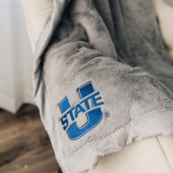 A Luxury Gray Colored Lush Saranoni Blanket. The soft blanket has a soft faux fur feeling. The blanket has the Utah State University Logo on it.