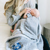 A mother holds her baby while holding a Luxury Toddler Gray Colored Lush Saranoni Blanket. The soft blanket is a toddler blanket or baby blanket and has a soft faux fur feeling. The blanket has the Utah State University Logo on it.