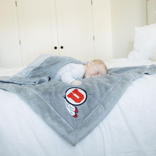 A baby lays on a Luxury Toddler Gray Colored Lush Saranoni Blanket. The soft blanket is a toddler blanket or baby blanket and has a soft faux fur feeling. The blanket has the University of Utah Logo on it.