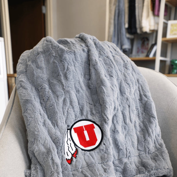 A Luxury Throw Gray Colored Patterned Faux Fur Saranoni Blanket. The soft blanket is a throw blanket and has a soft faux fur feeling. The blanket has a University of Utah Logo.