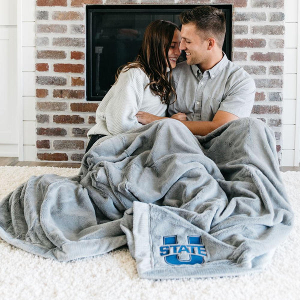 A husband and wife sit with a Luxury Extra Large Oversized Throw Gray Colored Lush Saranoni Blanket. The soft blanket has a soft faux fur feeling. The extra large blanket has the Utah State University Logo on it.