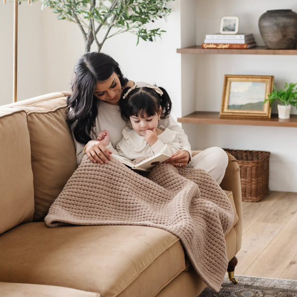 Mom reading book on couch with her daughter snuggled up with tan waffle knit blanket.