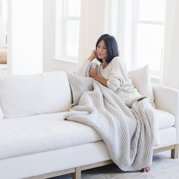 Woman sitting on couch snuggled up with a light gray waffle knit blanket. 